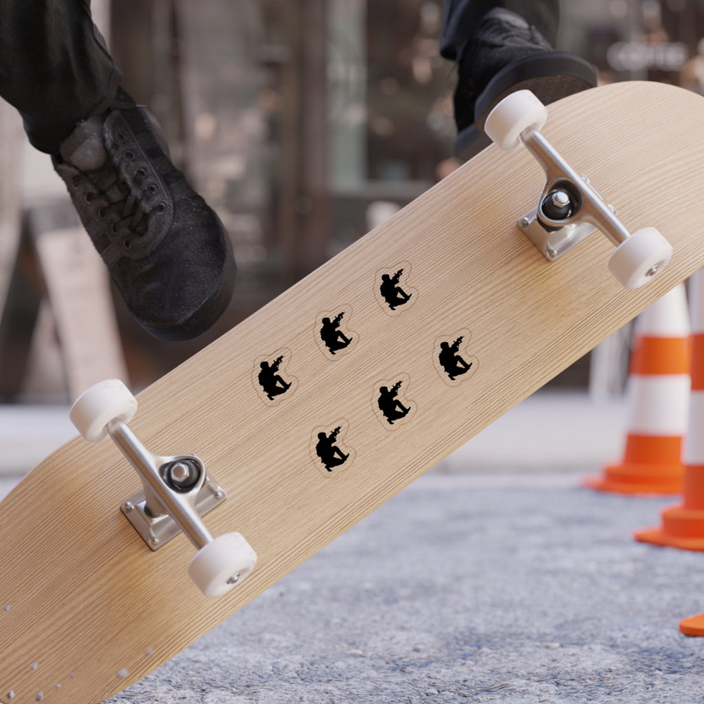 A close-up of a skateboard deck with six black skateboarder silhouettes arranged in a circle, photographed on an outdoor street with a person's feet and black sneakers visible at the top edge.